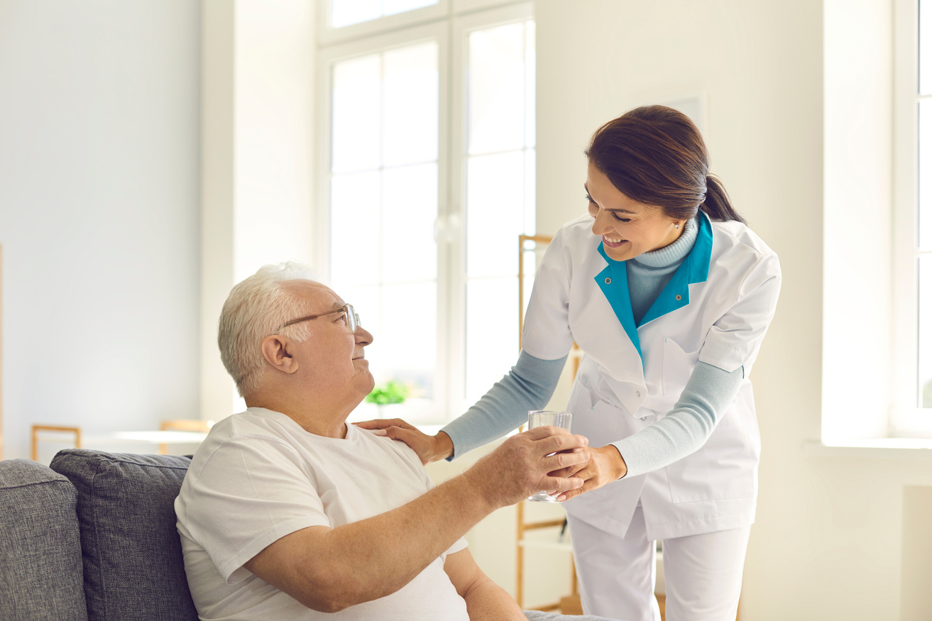Smiling Nurse Giving Glass of Water to Senior Man in Nursing Home or Assisted Living Facility